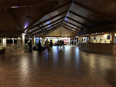 A spacious hotel lobby with a high wooden ceiling and tiled floor. Several people are gathered near a counter, possibly checking in or out. The lighting is warm and dim, creating a cozy atmosphere. There are decorative elements such as plants and wall art. The reception desk is staffed and well-lit, and there is a seating area visible in the background.
