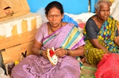 Two women are sitting on the ground, one wearing a purple and white patterned sari with colorful stripes and holding a decorative item made with flowers. The other woman is dressed in a green and blue checkered sari. They are seated in front of cardboard boxes and surrounded by a variety of colored fabrics and netting.