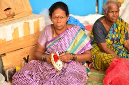 Two women are sitting on the ground, one wearing a purple and white patterned sari with colorful stripes and holding a decorative item made with flowers. The other woman is dressed in a green and blue checkered sari. They are seated in front of cardboard boxes and surrounded by a variety of colored fabrics and netting.