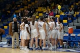A women's basketball team huddles together on the court, wearing white and blue uniforms with numbers. A coach and another individual stand nearby, providing guidance. Spectators are seated in the bleachers, some holding balloons, within a brightly lit indoor sports arena.