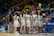 A women's basketball team huddles together on the court, wearing white and blue uniforms with numbers. A coach and another individual stand nearby, providing guidance. Spectators are seated in the bleachers, some holding balloons, within a brightly lit indoor sports arena.