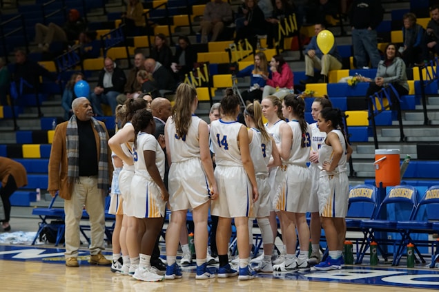 A women's basketball team huddles together on the court, wearing white and blue uniforms with numbers. A coach and another individual stand nearby, providing guidance. Spectators are seated in the bleachers, some holding balloons, within a brightly lit indoor sports arena.