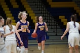 A group of young women are playing basketball indoors. Two players wearing purple uniforms with 'Bruins' are moving with one holding the basketball. They are facing players in white uniforms, with the gym's multicolored bleachers visible in the background.