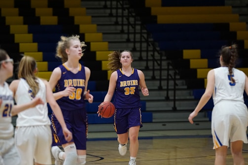 A group of young women are playing basketball indoors. Two players wearing purple uniforms with 'Bruins' are moving with one holding the basketball. They are facing players in white uniforms, with the gym's multicolored bleachers visible in the background.