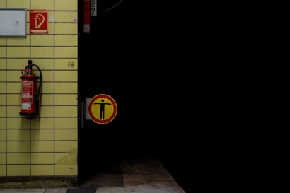 A tiled wall with a mounted fire extinguisher is partially visible alongside an open door leading into darkness. A yellow caution sign featuring a figure with outstretched arms is attached to the door.