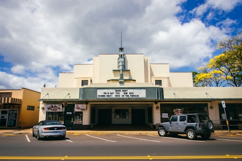 A vintage-style cinema with an art deco facade and marquee displaying movie titles. Two cars are parked in front of the building, and there is a tree with yellow flowers on the right. The sky is partly cloudy with patches of blue.