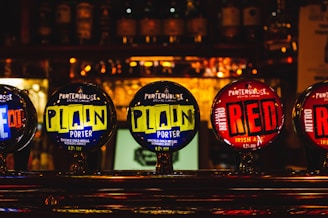 A close-up of beer taps at a bar featuring various labels, including 'Porterhouse Brewing Company'. The labels on the taps display vibrant colors with brand names 'Plain Porter' and 'Nitro Red Irish Ale'. Background is blurred with bottles visible on shelves.