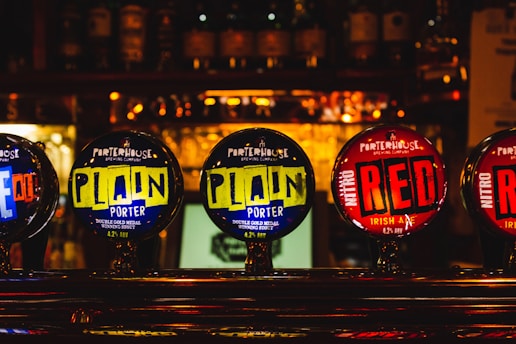 A close-up of beer taps at a bar featuring various labels, including 'Porterhouse Brewing Company'. The labels on the taps display vibrant colors with brand names 'Plain Porter' and 'Nitro Red Irish Ale'. Background is blurred with bottles visible on shelves.