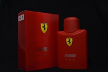 A bright red box and bottle with the Scuderia Ferrari logo prominently displayed. The box and bottle both feature the words 'Scuderia Ferrari' and 'Red' written in sleek silver text. The background is a plain dark color, which emphasizes the vivid red of the products.