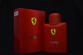 A bright red box and bottle with the Scuderia Ferrari logo prominently displayed. The box and bottle both feature the words 'Scuderia Ferrari' and 'Red' written in sleek silver text. The background is a plain dark color, which emphasizes the vivid red of the products.