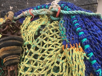 Various intertwined fishing nets with vibrant colors, including green, blue, and yellow ropes, are shown in a close-up. The image also features some rusted metal elements, such as chains and weights. The nets appear to be on a quay or dock, with a rough stone wall in the background.