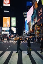 A bustling urban scene at night with people crossing a street filled with bright digital billboards and lights from surrounding buildings. The environment is lively with heavy pedestrian and vehicular traffic, featuring taxis and advertising signs for various brands.