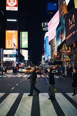 A bustling urban scene at night with people crossing a street filled with bright digital billboards and lights from surrounding buildings. The environment is lively with heavy pedestrian and vehicular traffic, featuring taxis and advertising signs for various brands.