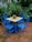 A set of four vibrant blue metal chairs and a matching square table with a mosaic-patterned surface are situated on a tiled patio. The table features a small, colorful potted plant with red flowers at the center. Lush green foliage surrounds the patio, creating a secluded and inviting atmosphere.