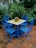 A set of four vibrant blue metal chairs and a matching square table with a mosaic-patterned surface are situated on a tiled patio. The table features a small, colorful potted plant with red flowers at the center. Lush green foliage surrounds the patio, creating a secluded and inviting atmosphere.