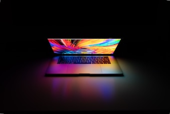 A laptop emits a vibrant, multicolored display while resting on a surface in a dimly lit environment. The screen features a swirling pattern of vivid colors, casting soft, colorful reflections on the surface.