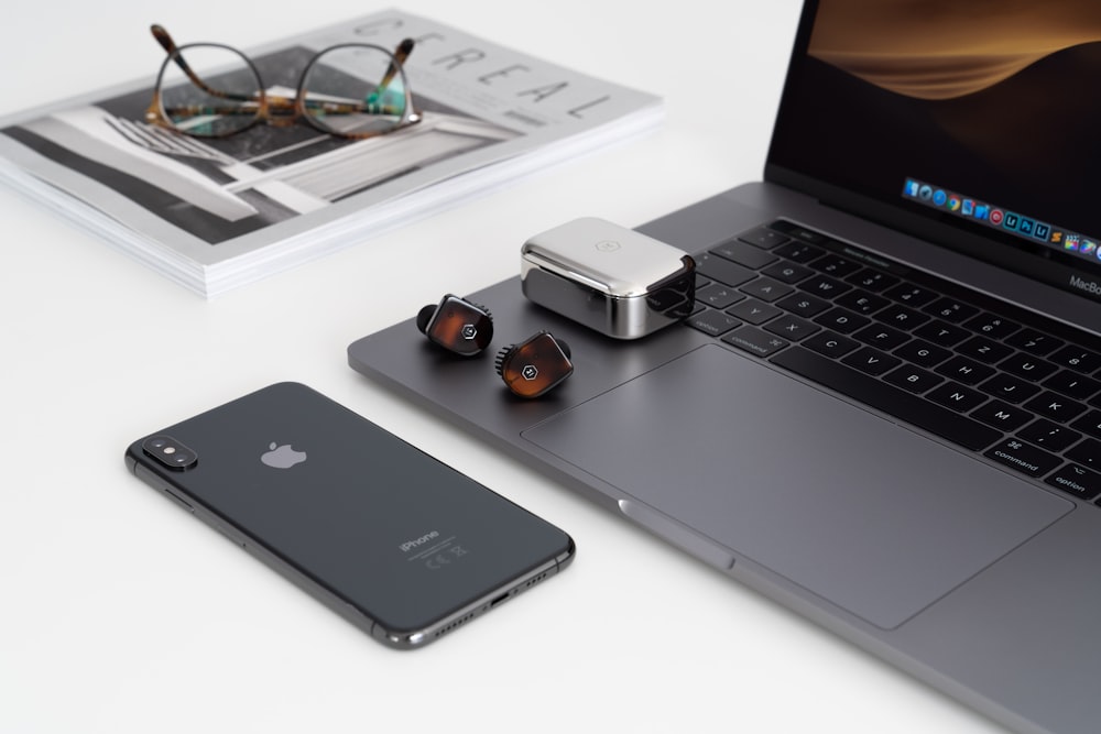 écouteurs Bluetooth sans fil avec étui argenté placé sur le bord du MacBook Pro devant l’iPhone X gris sidéral près des lunettes