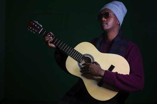 A person is playing a classical guitar while wearing a grey beanie, sunglasses, a maroon sweater, and a scarf. The focus is on the fingers and strings of the guitar, with a muted dark green background adding contrast to the image.