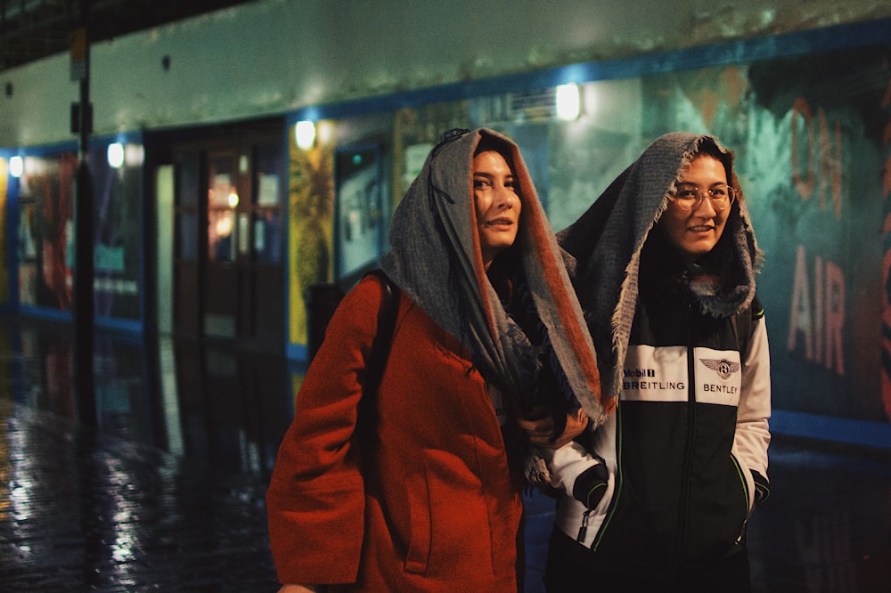 two smiling women sharing gray scarf covering heads