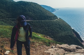 A person wearing a hat and outdoor clothing is hiking on a trail along a scenic coastal cliff. The background is filled with lush green hills and the vast blue expanse of the ocean.