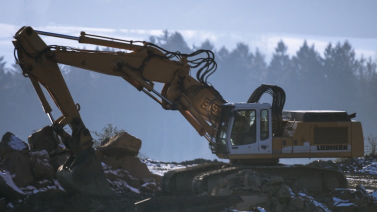 An excavator is stationed on rocky terrain with its arm extended. The background shows a blurred silhouette of trees under a blue sky, suggesting early morning or late afternoon lighting.