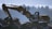 An excavator is stationed on rocky terrain with its arm extended. The background shows a blurred silhouette of trees under a blue sky, suggesting early morning or late afternoon lighting.