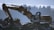 An excavator is stationed on rocky terrain with its arm extended. The background shows a blurred silhouette of trees under a blue sky, suggesting early morning or late afternoon lighting.