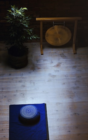 A serene meditation space with a wooden floor, featuring a blue mat and a meditation cushion placed on it. On the left, a potted plant adds a touch of nature. In the upper area, a gong hangs on a wooden stand against a brick wall, creating a calming atmosphere.