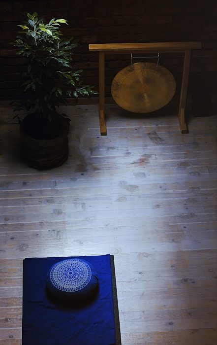 A serene meditation space with a wooden floor, featuring a blue mat and a meditation cushion placed on it. On the left, a potted plant adds a touch of nature. In the upper area, a gong hangs on a wooden stand against a brick wall, creating a calming atmosphere.