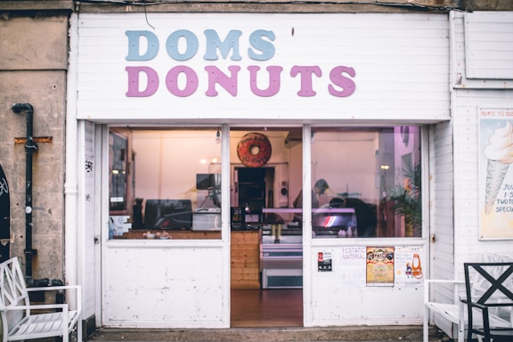 A small donut shop with a front sign that reads 'DOMS DONUTS' in pastel colors. The storefront has a glass window and door, allowing a view of the interior which features a wooden counter and some equipment. The exterior walls are white, and there are posters and chairs outside.