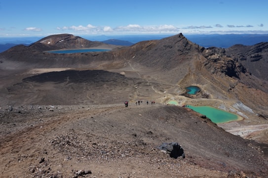 A barren volcanic landscape featuring rocky terrain and several distinctively colored lakes. A group of hikers traverses a trail in the foreground, with rugged mountains and rolling hills in the distance.