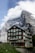 A rustic, multi-story chalet-style building is set against a dramatic mountainous backdrop. The building features a brown and beige facade with green shutters and balconies, and 'DESALPES' is prominently displayed on its front. The towering mountain behind is partly covered in snow and partially shrouded in clouds, creating a stunning contrast with the architecture.