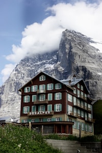 A rustic, multi-story chalet-style building is set against a dramatic mountainous backdrop. The building features a brown and beige facade with green shutters and balconies, and 'DESALPES' is prominently displayed on its front. The towering mountain behind is partly covered in snow and partially shrouded in clouds, creating a stunning contrast with the architecture.