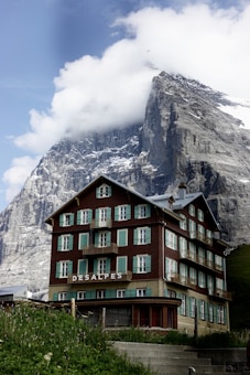A rustic, multi-story chalet-style building is set against a dramatic mountainous backdrop. The building features a brown and beige facade with green shutters and balconies, and 'DESALPES' is prominently displayed on its front. The towering mountain behind is partly covered in snow and partially shrouded in clouds, creating a stunning contrast with the architecture.