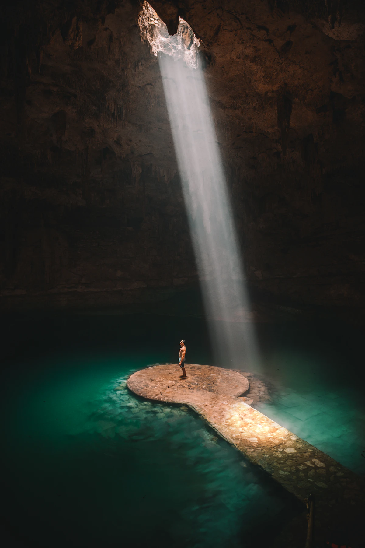 A shaft of daylight falling through the roof of a Yucatán cenote onto still turquoise water below.