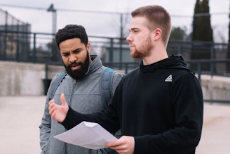 man in black hoodie holding white printer paper and man in grey zip-up jacket during daytime