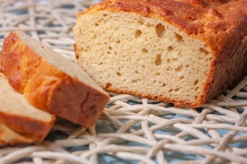 A loaf of bread is placed on a woven wicker surface, with two slices partially visible in the foreground. The bread has a golden-brown crust and a soft, airy texture inside.