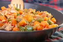A close-up of a colorful stir-fry dish in a pan, featuring diced carrots, green bell peppers, chicken pieces, and onions. The dish appears vibrant and freshly cooked, with a wooden spatula visible in the background.