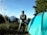 A person in outdoor clothing stands next to a blue tent, holding a tripod. In the background, a mountain can be seen under a clear blue sky, surrounded by green trees and foliage. The scene suggests a camping trip with natural surroundings.