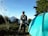 A person in outdoor clothing stands next to a blue tent, holding a tripod. In the background, a mountain can be seen under a clear blue sky, surrounded by green trees and foliage. The scene suggests a camping trip with natural surroundings.