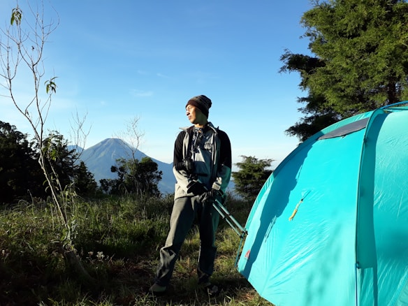 A person in outdoor clothing stands next to a blue tent, holding a tripod. In the background, a mountain can be seen under a clear blue sky, surrounded by green trees and foliage. The scene suggests a camping trip with natural surroundings.
