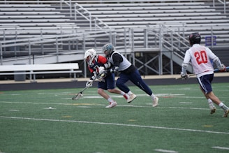 Three lacrosse players in motion on a field, focusing on one player in a red jersey and helmet being closely pursued by an opponent. The background features empty bleachers, emphasizing an outdoor sports setting.