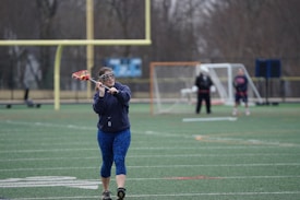 A person in sports attire is practicing lacrosse on a field. They are holding a lacrosse stick and wearing protective eyewear. In the background, there are some goalposts and other people involved in a sports activity.