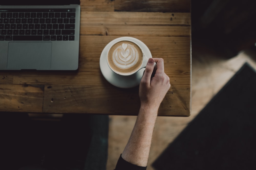 person holding cup of coffee on table beside laptop computer