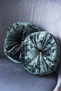 Two round velvet cushions with a subtle sheen are placed against a matching velvet textured background. Each cushion features a button at the center, adding a touch of elegance.