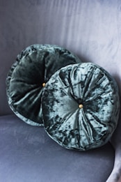 Two round velvet cushions with a subtle sheen are placed against a matching velvet textured background. Each cushion features a button at the center, adding a touch of elegance.