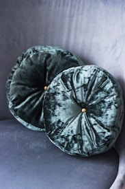 Two round velvet cushions with a subtle sheen are placed against a matching velvet textured background. Each cushion features a button at the center, adding a touch of elegance.
