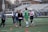 A group of people are on a sports field engaging in a casual interaction. Several individuals are holding lacrosse sticks, suggesting they are players. The scene appears to capture a practice or friendly game setting, with casual and sport-specific attire worn by the participants. The background consists of bleachers, indicating this is part of a larger sports facility.