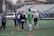 A group of people are on a sports field engaging in a casual interaction. Several individuals are holding lacrosse sticks, suggesting they are players. The scene appears to capture a practice or friendly game setting, with casual and sport-specific attire worn by the participants. The background consists of bleachers, indicating this is part of a larger sports facility.