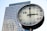 A large, modern clock is prominently displayed in the foreground, with a high-rise office building featuring numerous windows in the background. The clock face is white with black hour and minute markers, and a red second hand.